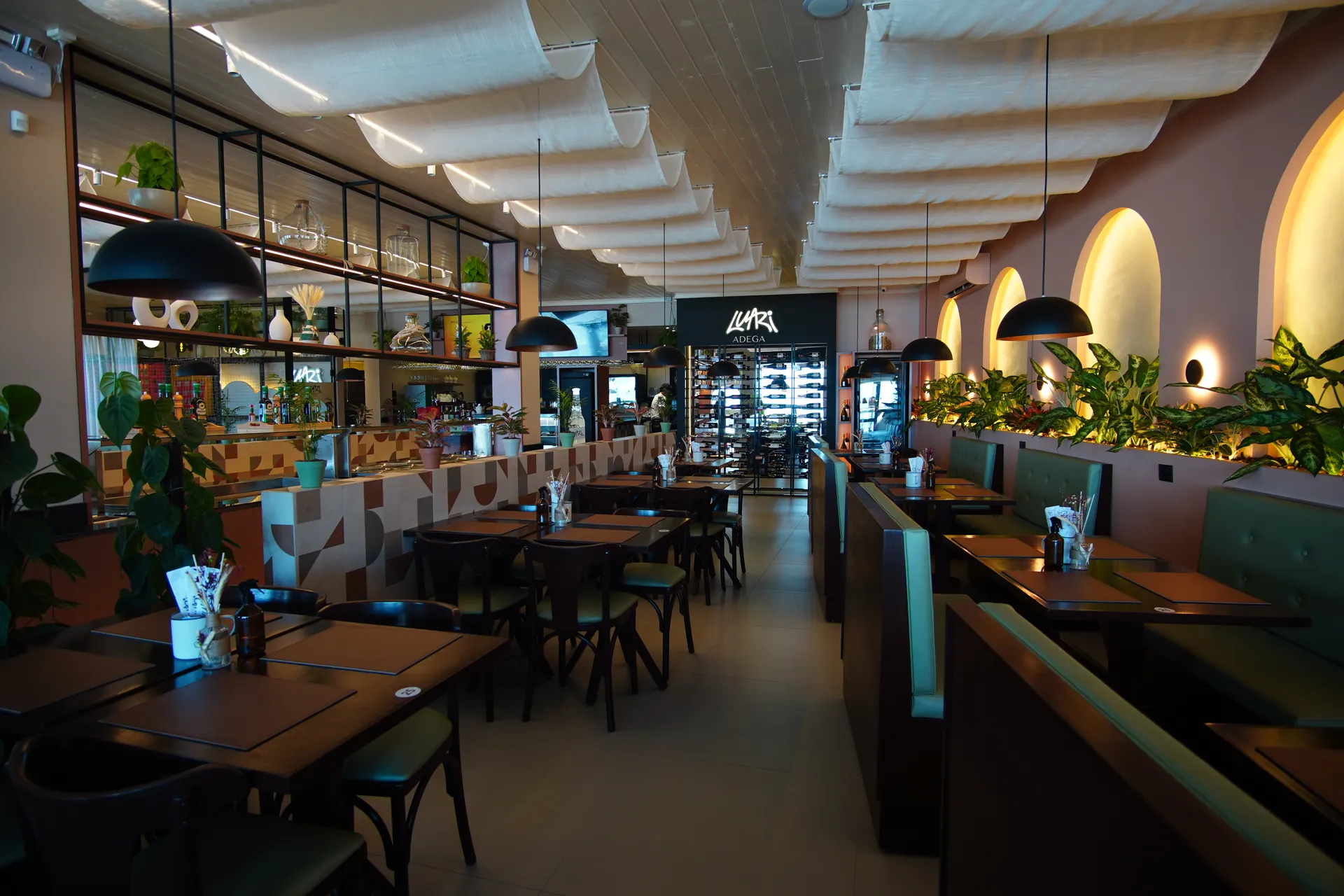 Salão do restaurante Luari, com mesas de madeira, sofás verdes, jardineiras iluminadas e forro de tecido